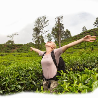 Active caucasian blonde woman, arms raised to sky, enjoing fresh air and pristine nature while tracking among tea plantaitons near Ella, Sri Lanka. Bacpecking outdoors tourist adventure.
